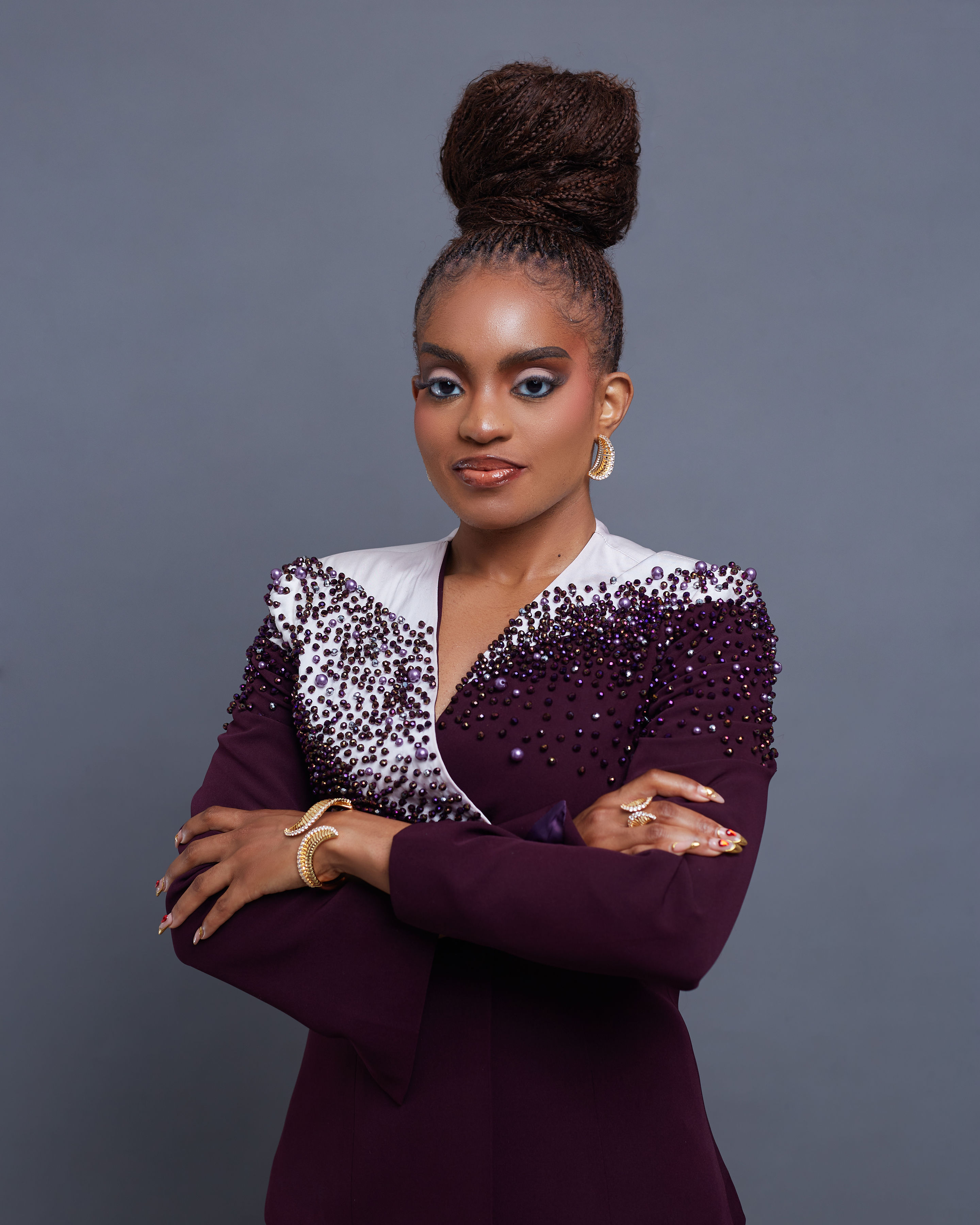 Portrait of founder Adaobi Onyekaba with arms crossed, wearing the Chikamso Suit — a deep burgundy tailored jacket with ombré crystal and pearl beadwork migrating from a white satin lapel across a structured shoulder.