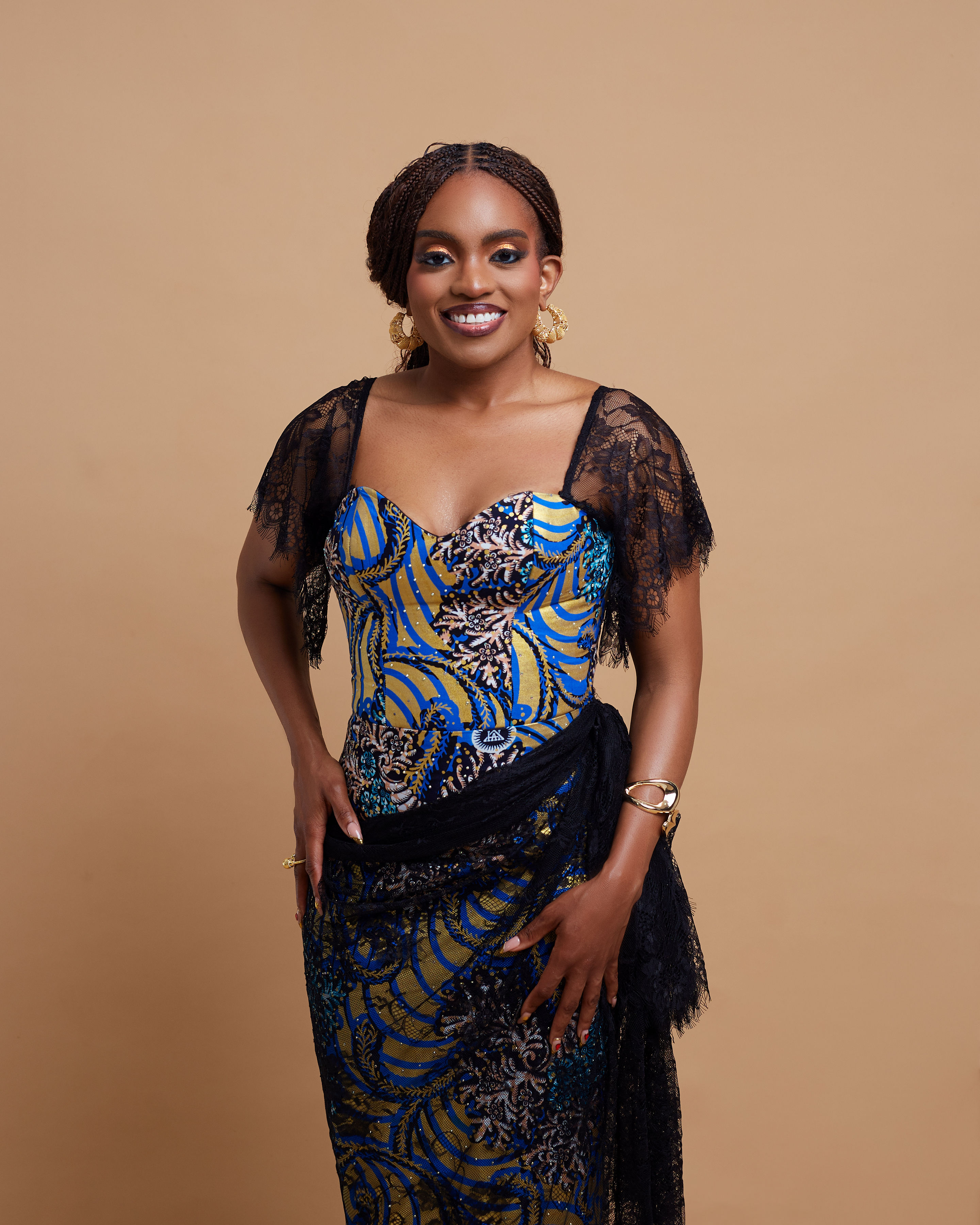 Full-length view of the Mmesoma gown on founder Adaobi Onyekaba, its black Chantilly lace overlay and matching wrapper draping knotted at the hip, photographed against a warm tan backdrop.