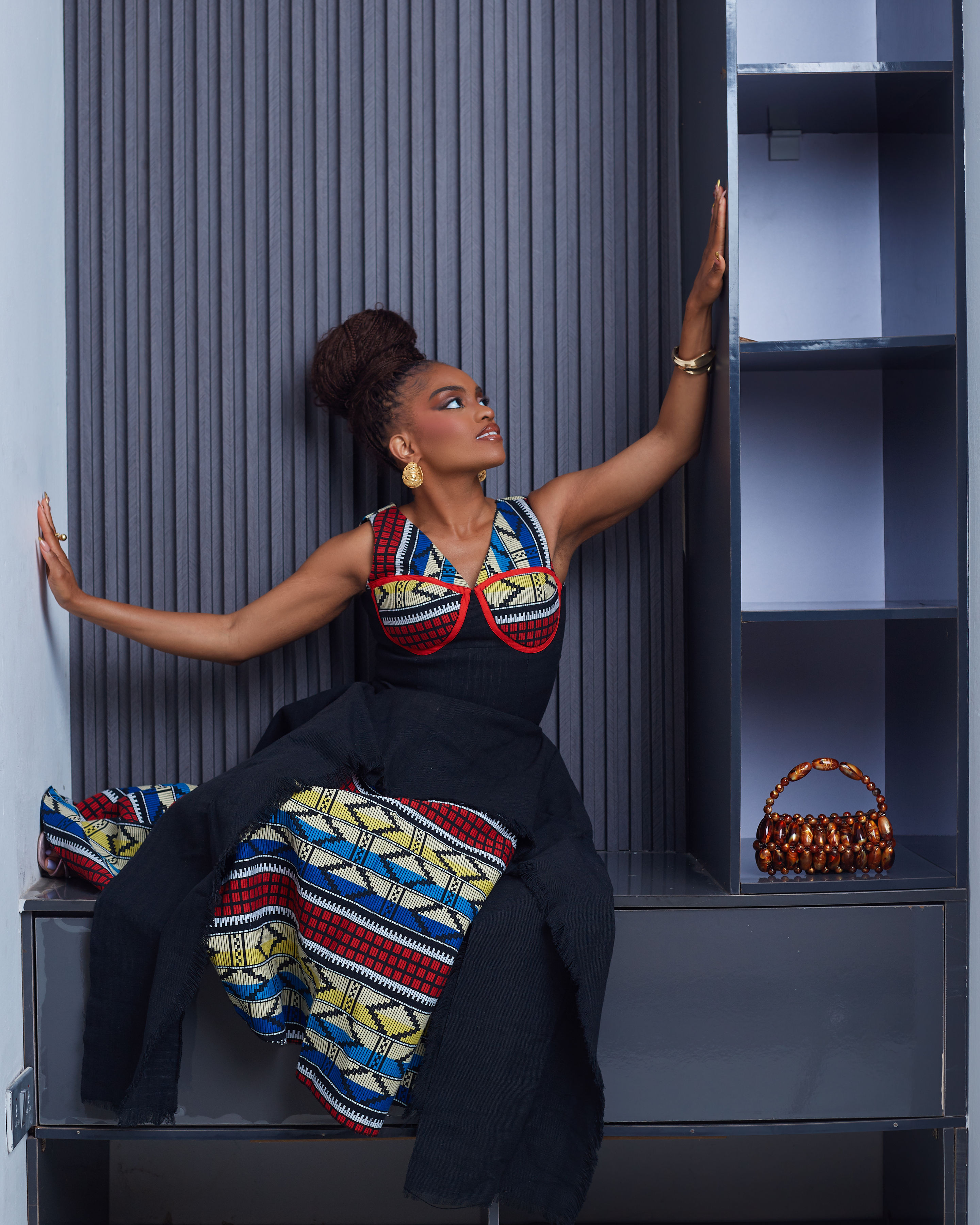 Standing full-length view of the Ifeyinwa dress, the woven Akwete skirt visible beneath the fringed black Aso-Oke tiers, founder Adaobi Onyekaba holding an amber beaded bag.